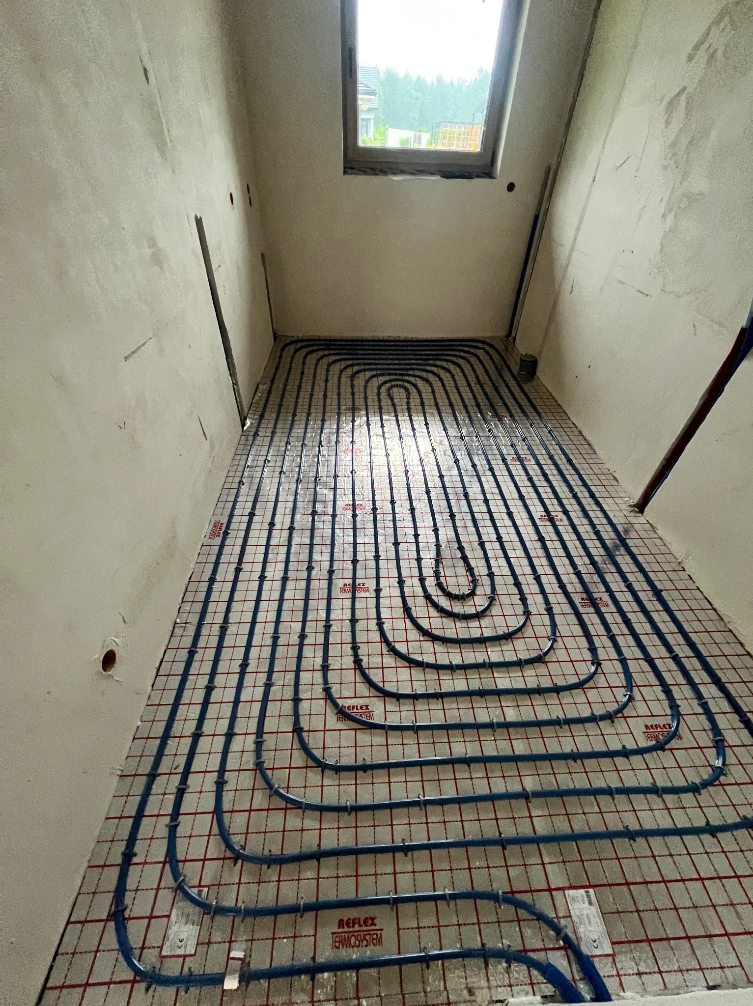 Niebieskie rury ogrzewania podłogowego ułożone spiralnie na czerwonej macie w surowym wnętrzu. Widok z góry na instalację przed wylaniem posadzki.