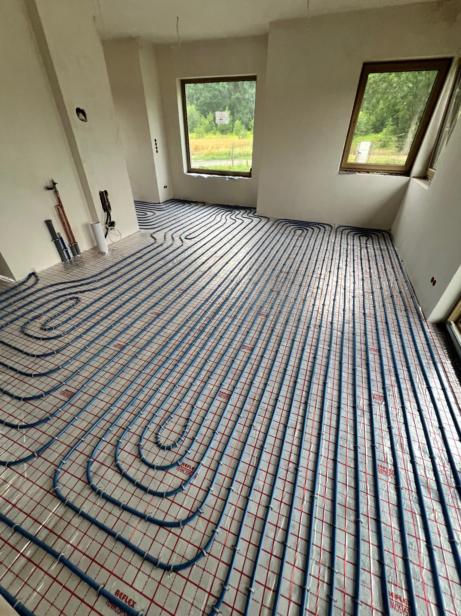 Niebieskie rury ogrzewania podłogowego ułożone równolegle na folii z czerwoną siatką w jasnym pokoju z dwoma oknami. Widoczne narzędzia instalacyjne przy ścianie.