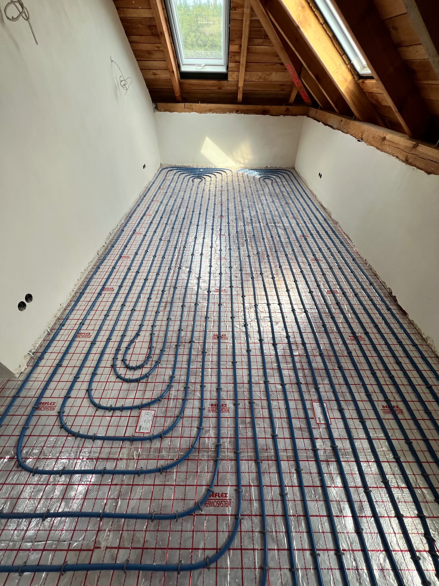 Układanie niebieskich rur ogrzewania podłogowego na folii w poddaszu ze skosami i oknem dachowym. Widoczne mocowania rur. Etap przed wylewką.