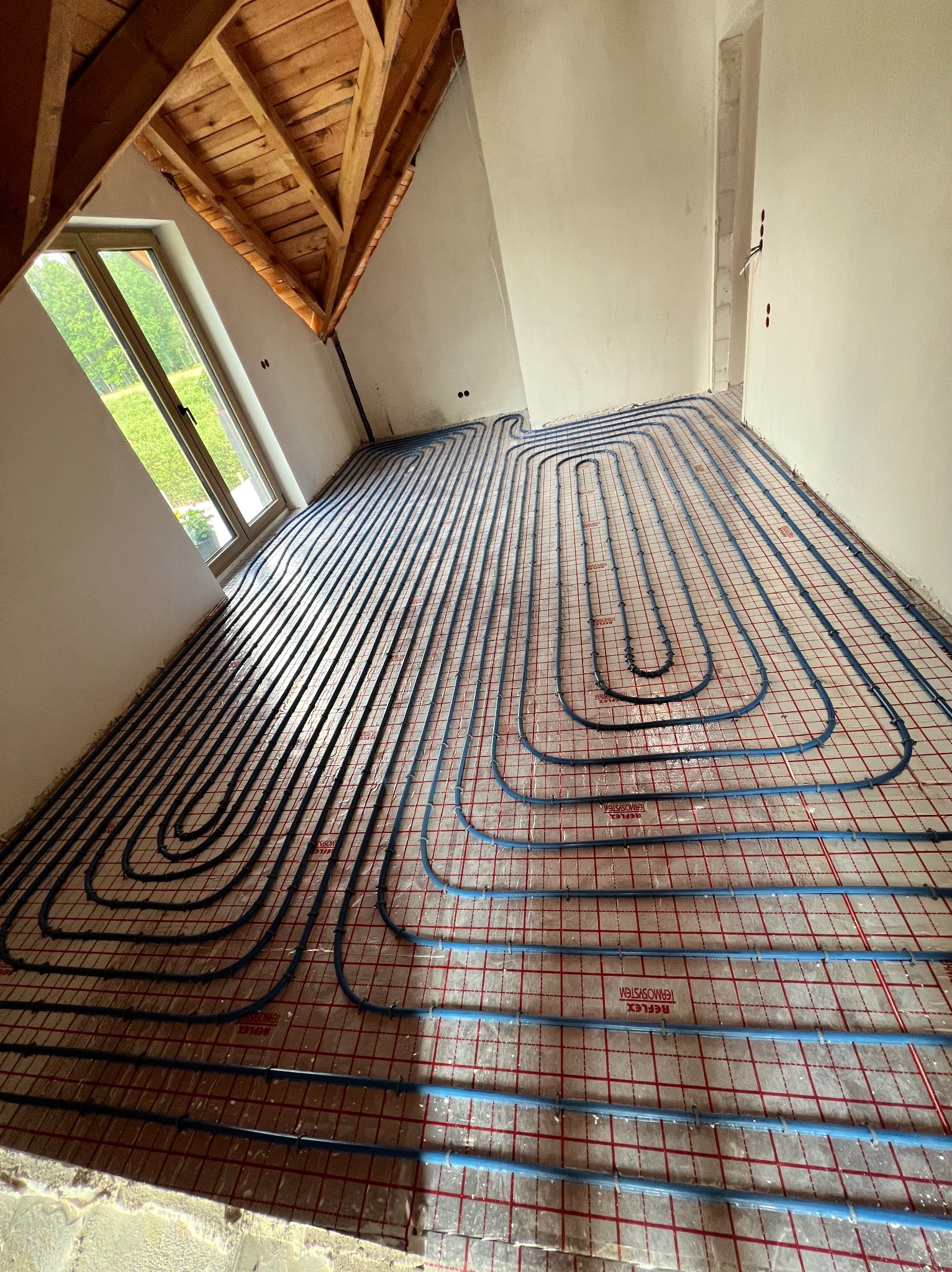 Ułożone, niebieskie rury ogrzewania podłogowego na czerwonej siatce w jasnym pokoju na poddaszu z oknem. Widoczne drewniane elementy konstrukcji dachu.