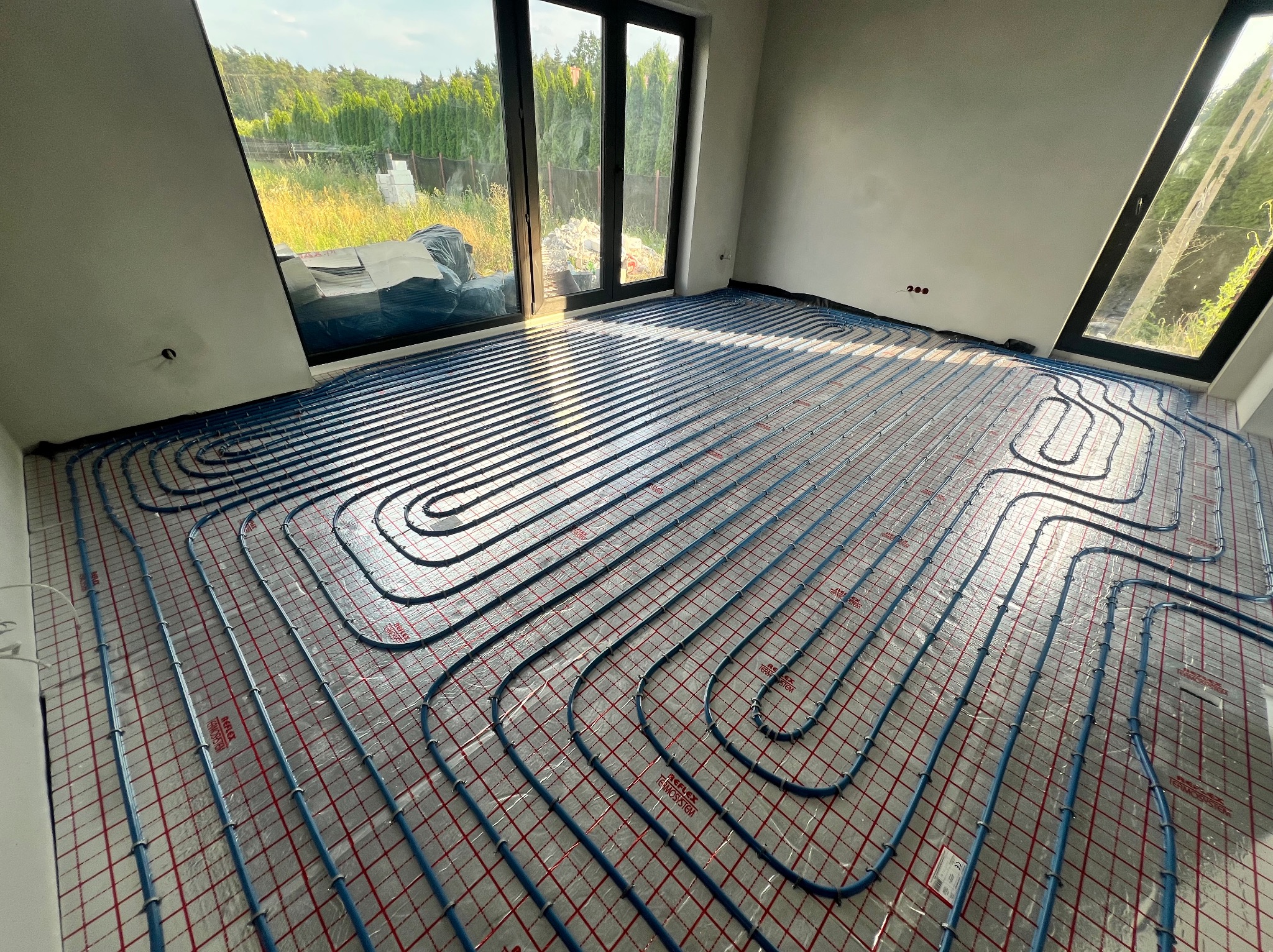 Błękitne rury ogrzewania podłogowego ułożone na folii z czerwoną kratką w jasnym, surowym wnętrzu z dużymi oknami, przez które widać zieleń.