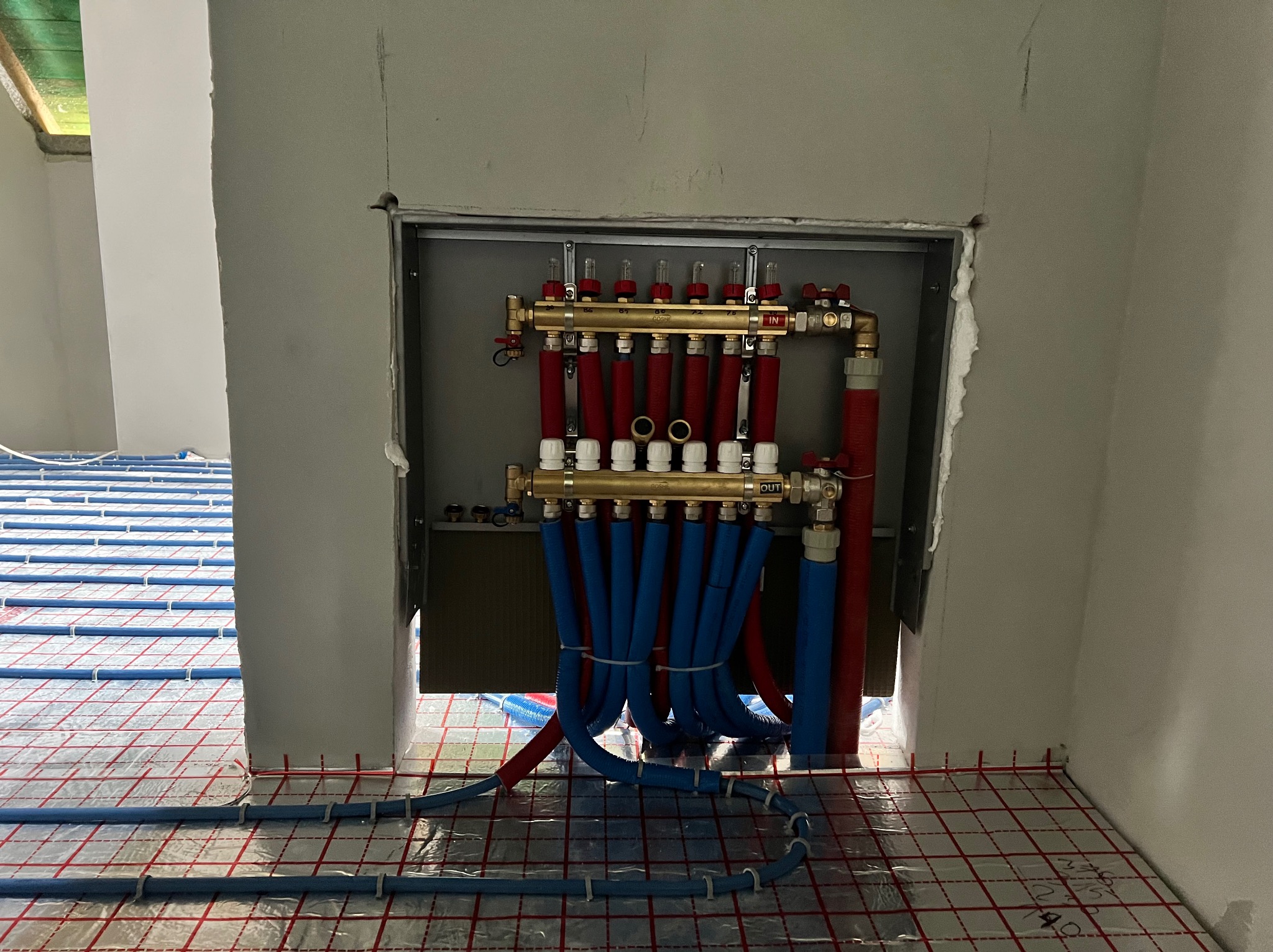 Rozdzielacz ogrzewania podłogowego z rurami w kolorze niebieskim i czerwonym, zamontowany w szarej wnęce ściany. Widoczna instalacja na podłodze z folią.