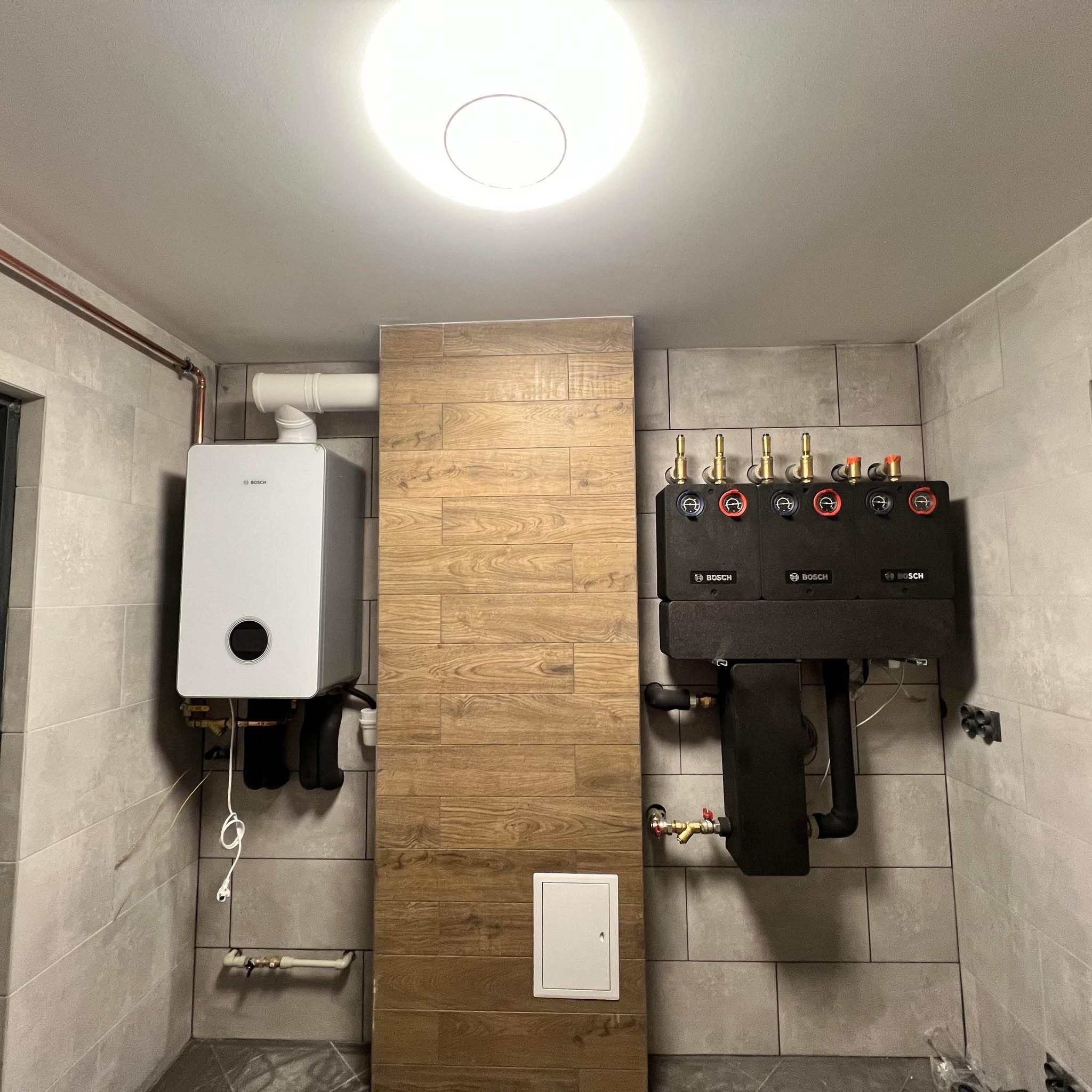 Nowoczesna kotłownia z białym piecem gazowym Bosch i czarnym rozdzielaczem na tle szarych płytek i drewnopodobnej wstawki. Widoczne rury i elementy instalacji.