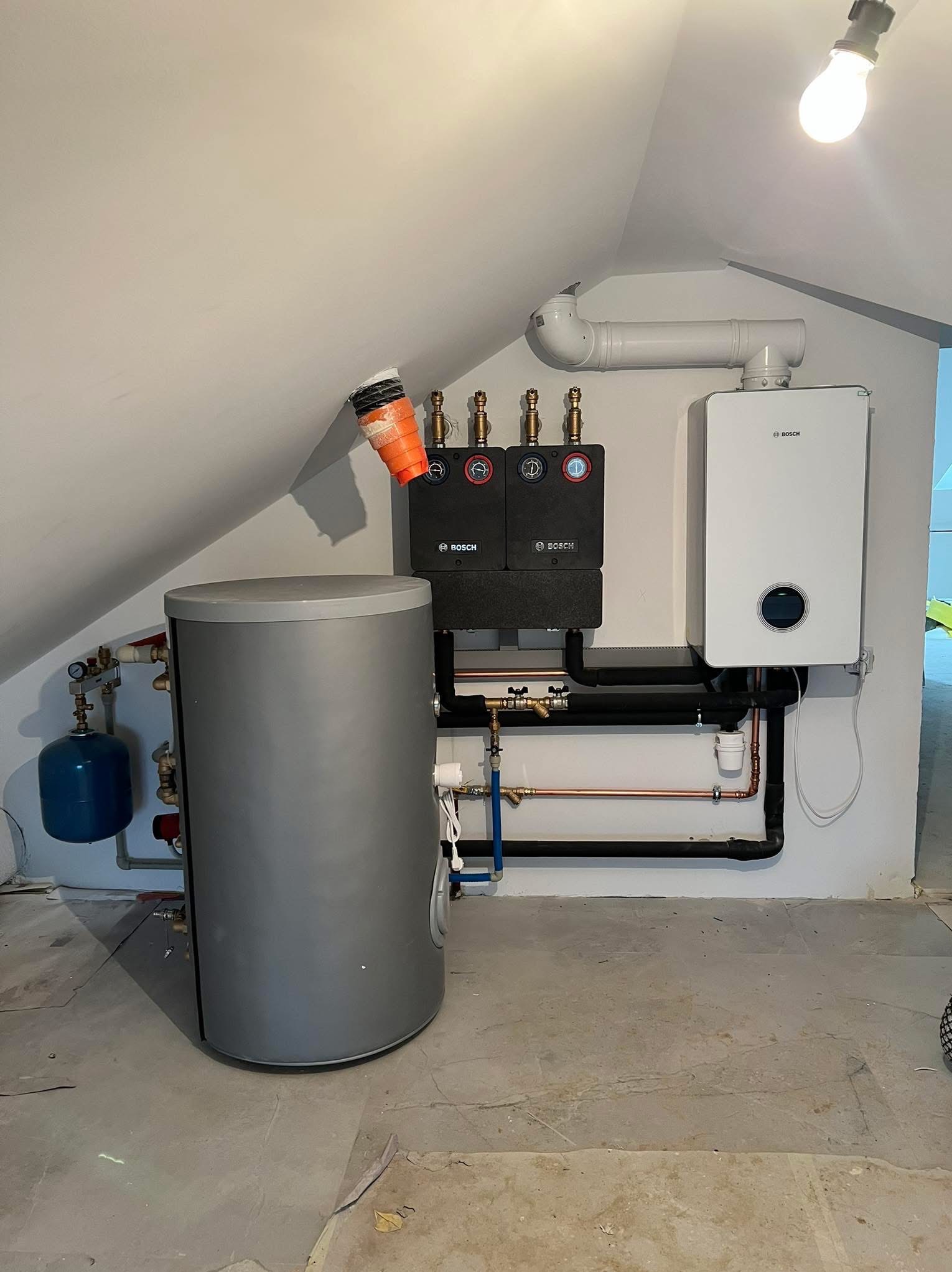 Nowoczesna kotłownia w skosie poddasza: szary zasobnik, biały piec gazowy Bosch, czarne rozdzielacze i rury. Widoczne elementy instalacji i oświetlenie.