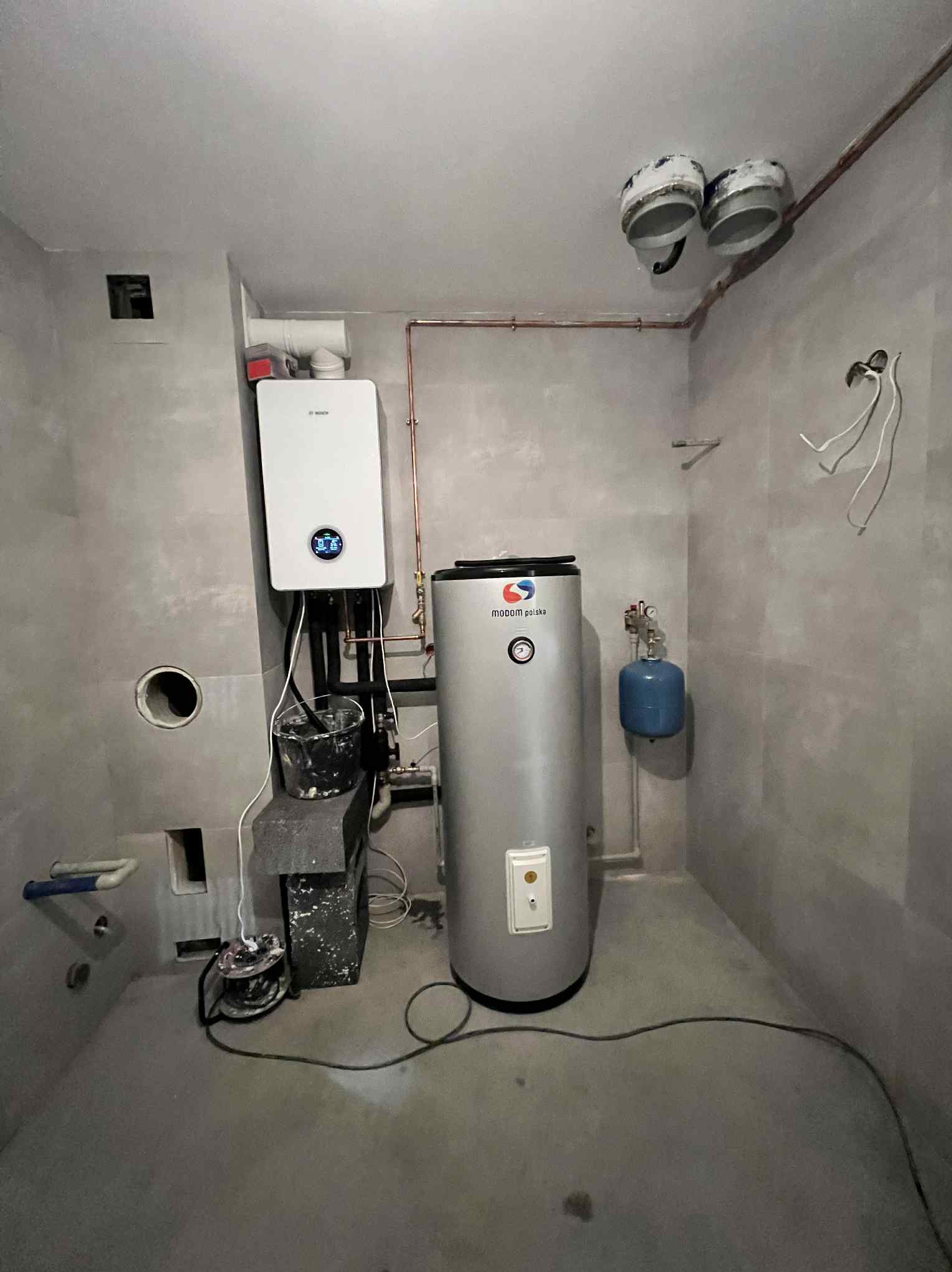 Nowoczesny piec gazowy Bosch i srebrny zasobnik Modom Polska w surowym pomieszczeniu z instalacją miedzianą. Widoczne elementy instalacji i akcesoria montażowe.