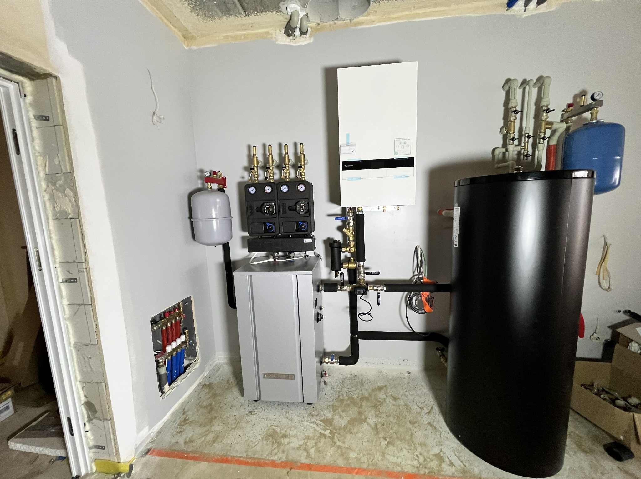 Nowoczesna kotłownia w trakcie montażu: biały piec gazowy, czarny zbiornik, szara szafka sterownicza i instalacja hydrauliczna w surowym pomieszczeniu.
