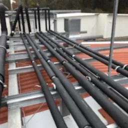 Izolowane rury wentylacyjne układane na metalowej konstrukcji wsporczej na dachu budynku, widoczne elementy systemu wentylacji zewnętrznej.