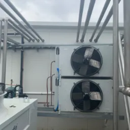 Dwa agregaty chłodnicze z wentylatorami na zewnątrz budynku, otoczone rurami instalacyjnymi, widoczne logo Thermofin na urządzeniu obok.