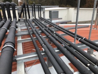 Izolowane rury wentylacyjne układane na metalowej konstrukcji wsporczej na dachu budynku, widoczne elementy systemu wentylacji zewnętrznej.