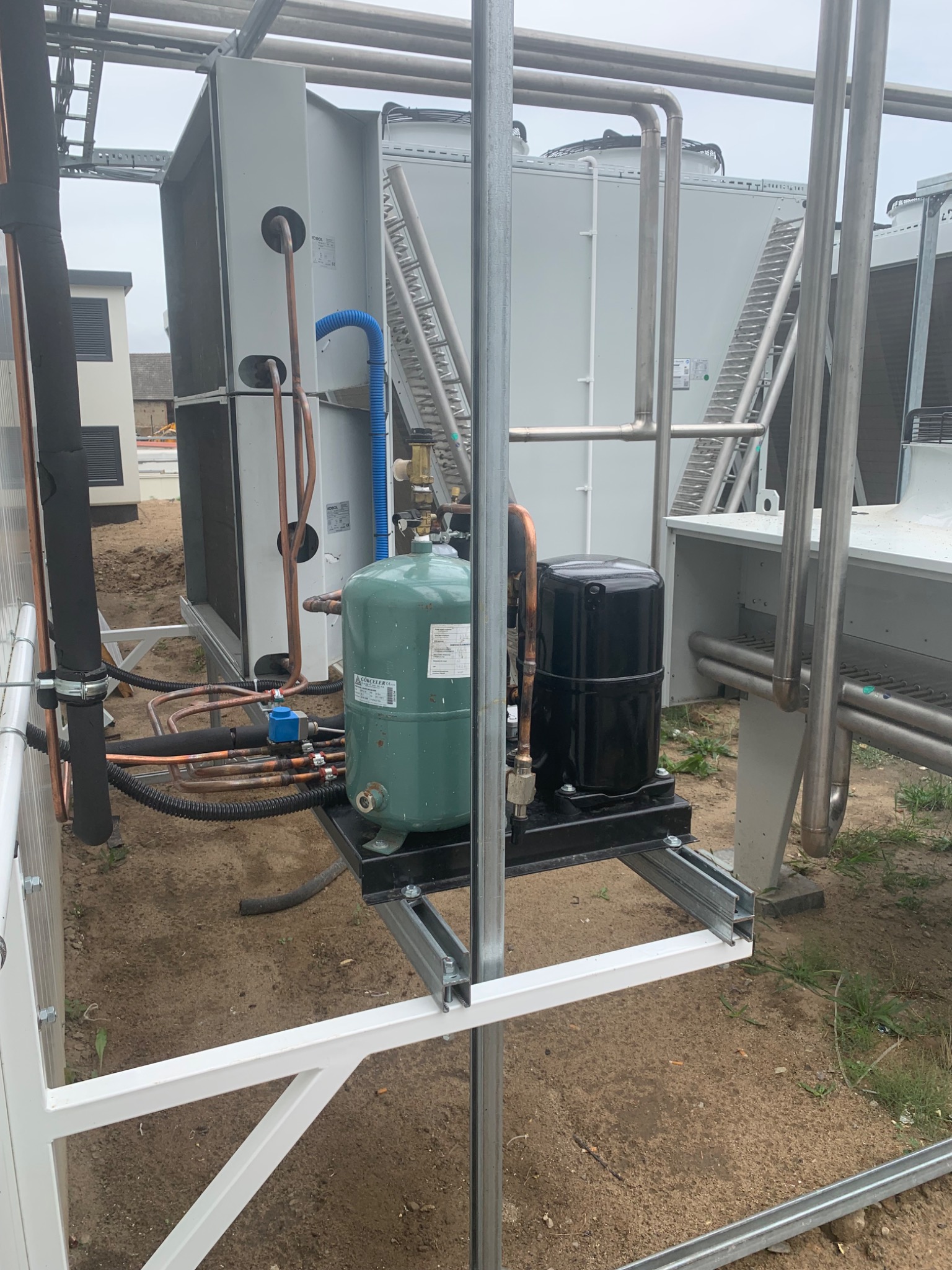 Instalacja chłodnicza na zewnątrz budynku, z widocznymi elementami: zbiornik ciśnieniowy w kolorze zielonym, czarny kompresor, rury miedziane, wentylatory chłodnicze oraz konstrukcja wsporcza.