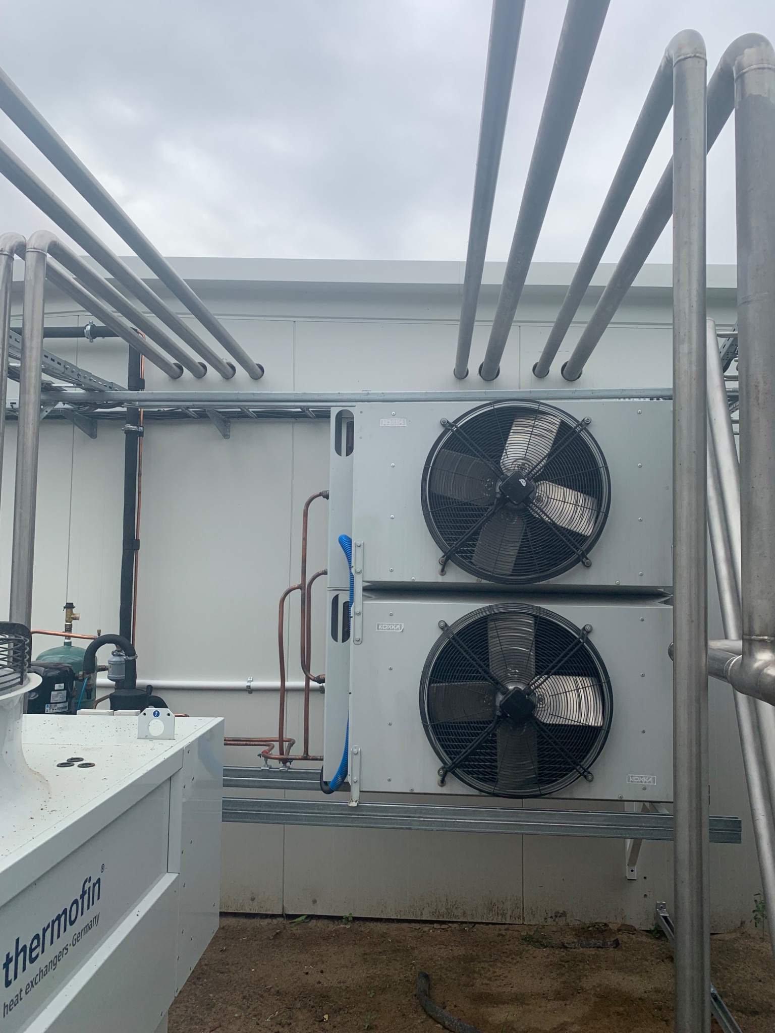 Dwa agregaty chłodnicze z wentylatorami na zewnątrz budynku, otoczone rurami instalacyjnymi, widoczne logo Thermofin na urządzeniu obok.