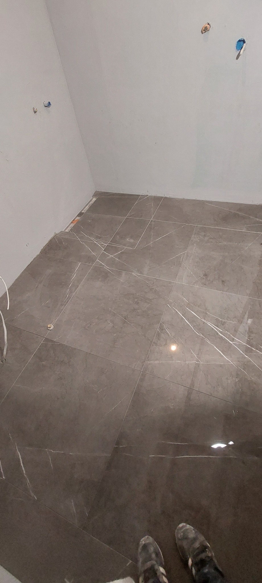 Ułożone, błyszczące, szare płytki podłogowe z białymi żyłkami w trakcie remontu łazienki, widoczne wystające kable i rury ze ściany oraz fragment butów.