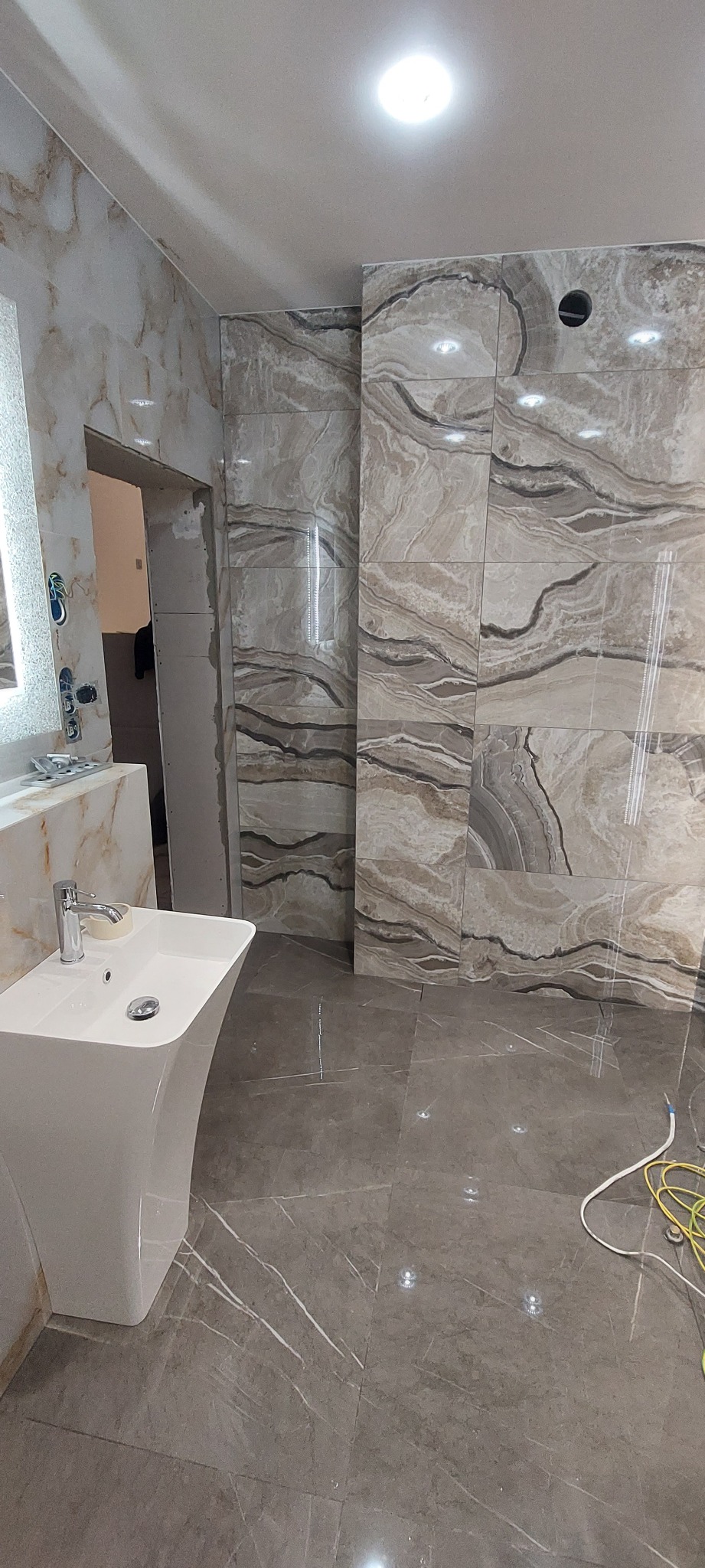Łazienka w trakcie wykańczania z marmurowymi płytkami na ścianach i podłodze, widoczna umywalka na postumencie, wystające kable i niedokończone obramowanie drzwi.