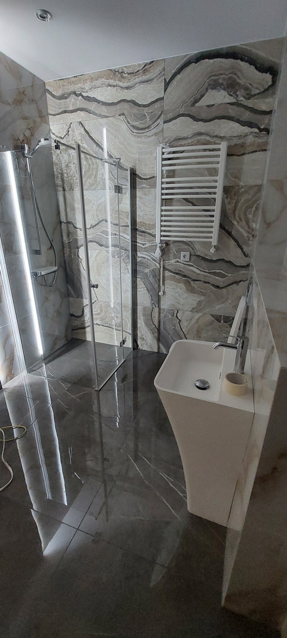 Nowoczesna łazienka z kabiną prysznicową ze szklanymi drzwiami, grzejnikiem drabinkowym i umywalką na postumencie, wykończona płytkami imitującymi marmur z wyraźnym wzorem żyłkowania, na podłodze...