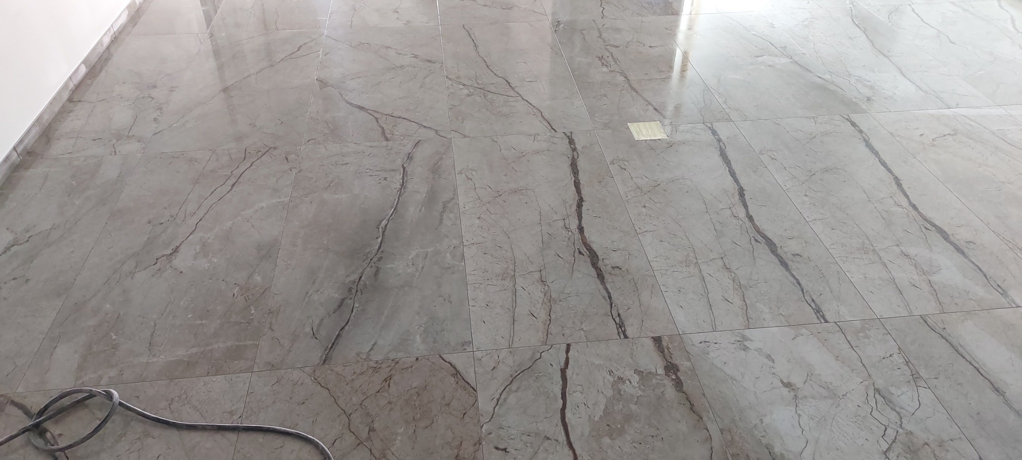 Ułożone płytki podłogowe imitujące marmur w odcieniach szarości z ciemniejszymi żyłami, widoczny kabel urządzenia w lewym dolnym rogu i fragment białej ściany.