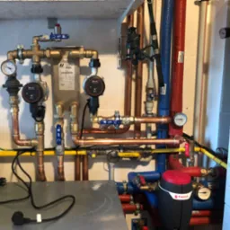Rozbudowana instalacja hydrauliczna i grzewcza w pomieszczeniu technicznym, z widocznymi miedzianymi rurami, pompami obiegowymi Keller, zaworami, manometrami, zbiornikiem Flamco i przewodami...