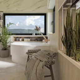 Jasna łazienka w Przemyślu z okrągłą wanną pod oknem z widokiem na góry, roślinami doniczkowymi i drewnianym stołkiem z narzuconym kocem.