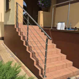 Zewnętrzne schody z terakoty z nowoczesną, minimalistyczną balustradą ze stali nierdzewnej, widok od dołu, z elementami ogrodu i fragmentem elewacji budynku w tle.