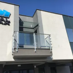 Zewnętrzny widok budynku z balkonem zabezpieczonym szklaną balustradą z metalowymi słupkami, nad wejściem daszek ze szkła, na ścianie logo firmy Metal Max.