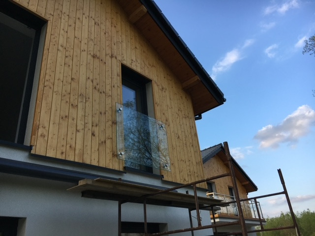 Surowy stan domu szkieletowego z drewnianą elewacją i zamontowanymi szklanymi barierkami balkonowymi, widoczne rusztowania.