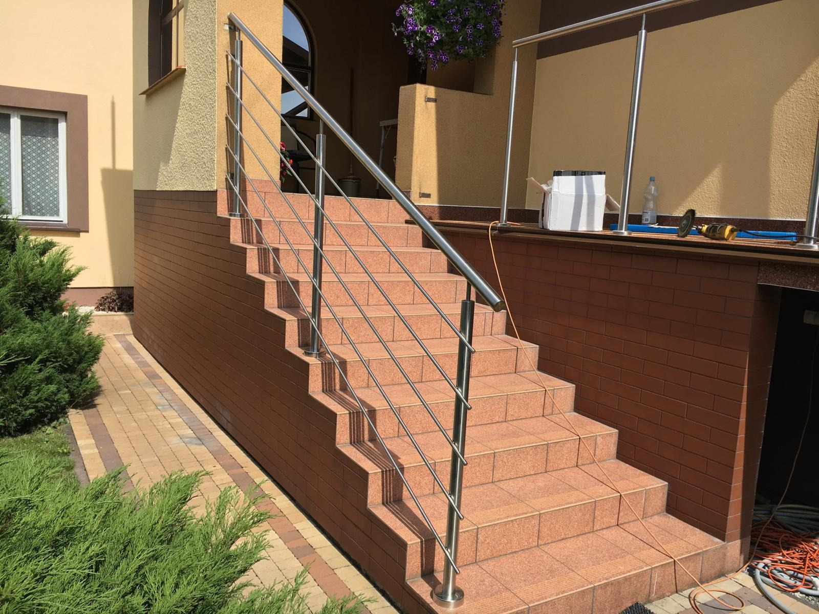 Zewnętrzne schody z terakoty z nowoczesną, minimalistyczną balustradą ze stali nierdzewnej, widok od dołu, z elementami ogrodu i fragmentem elewacji budynku w tle.