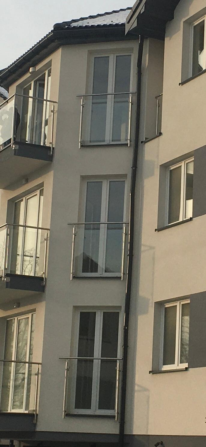 Fasada budynku wielorodzinnego z balkonami zabezpieczonymi szklanymi balustradami z metalowymi poręczami, widok z dołu na kilka kondygnacji.