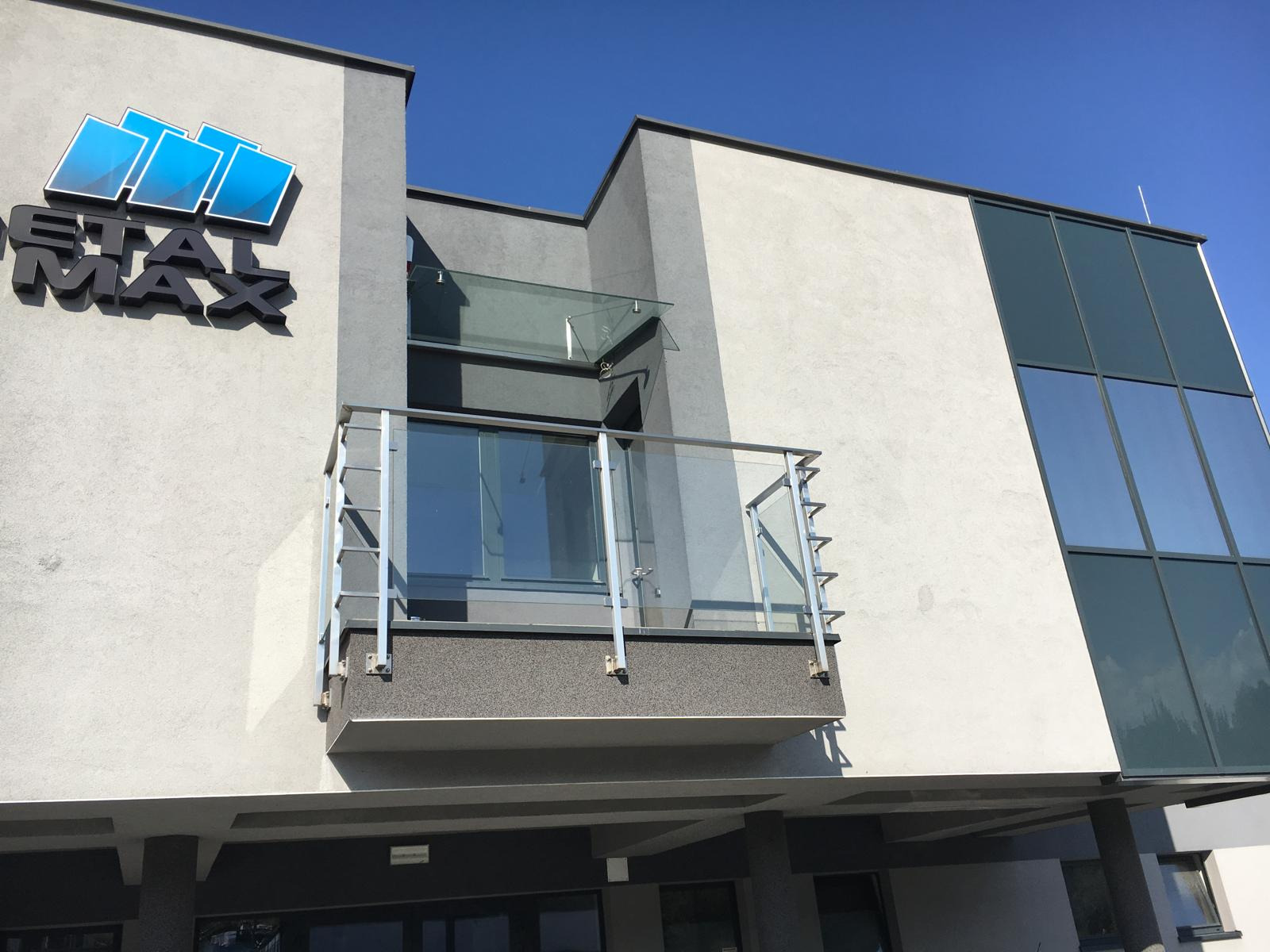 Zewnętrzny widok budynku z balkonem zabezpieczonym szklaną balustradą z metalowymi słupkami, nad wejściem daszek ze szkła, na ścianie logo firmy Metal Max.