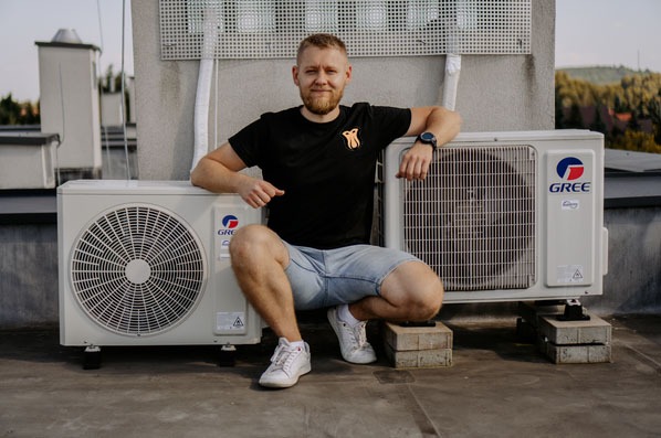 Mężczyzna w czarnej koszulce i jeansowych szortach kuca na betonowych bloczkach obok dwóch jednostek zewnętrznych klimatyzacji marki Gree na dachu budynku z widokiem na zalesione wzgórza w tle.