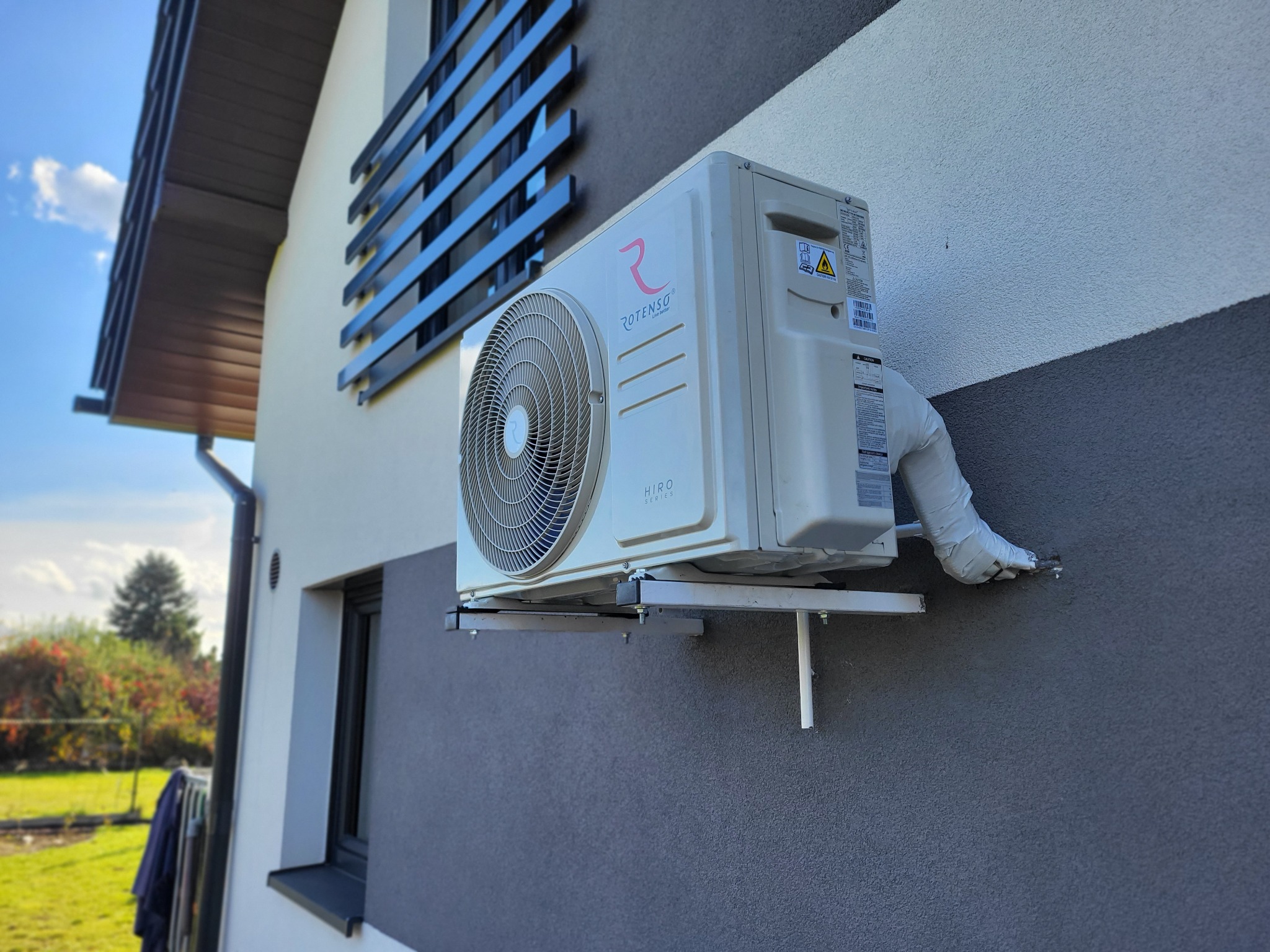 Biała jednostka zewnętrzna klimatyzatora Rotenso Hiro Series zamontowana na ścianie domu z szarą elewacją, widoczne rury odprowadzające skropliny owinięte białą taśmą izolacyjną.