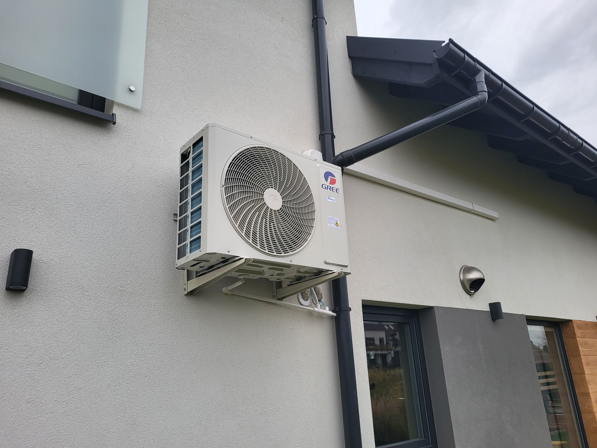 Biała jednostka zewnętrzna klimatyzacji marki Gree zamontowana na elewacji domu z widocznymi elementami instalacji odprowadzającej skropliny i rurami spustowymi.
