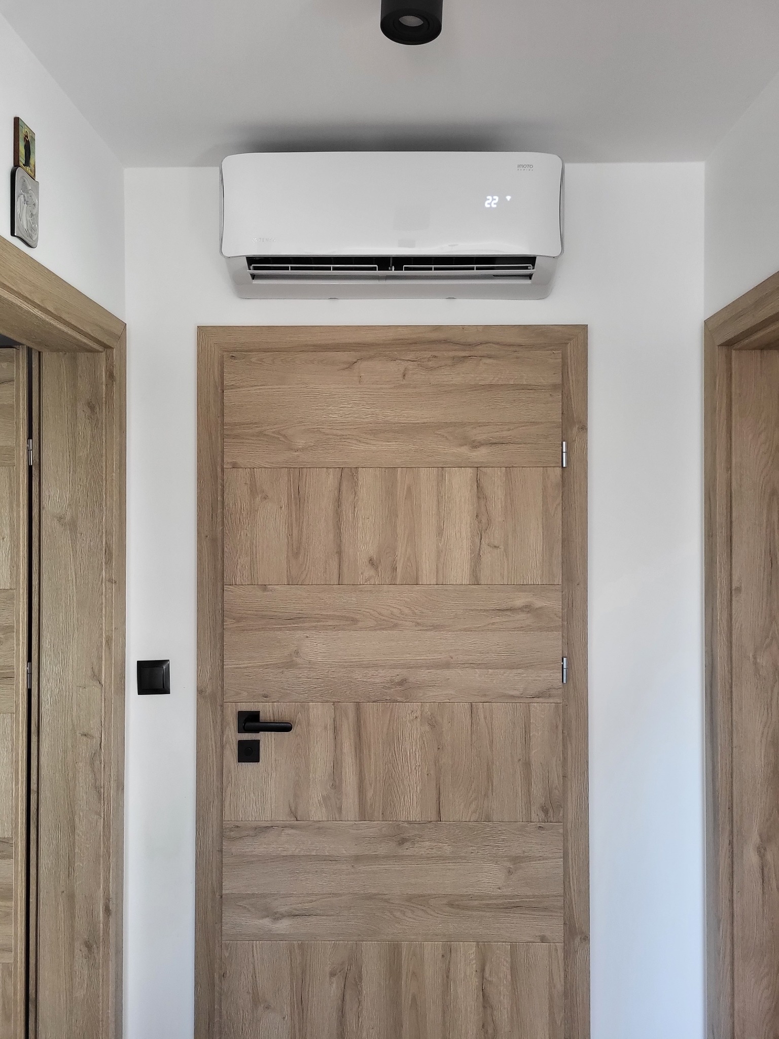 Biała jednostka klimatyzacyjna zamontowana nad drewnianymi drzwiami w nowoczesnym wnętrzu z białymi ścianami i czarnym oświetleniem punktowym.