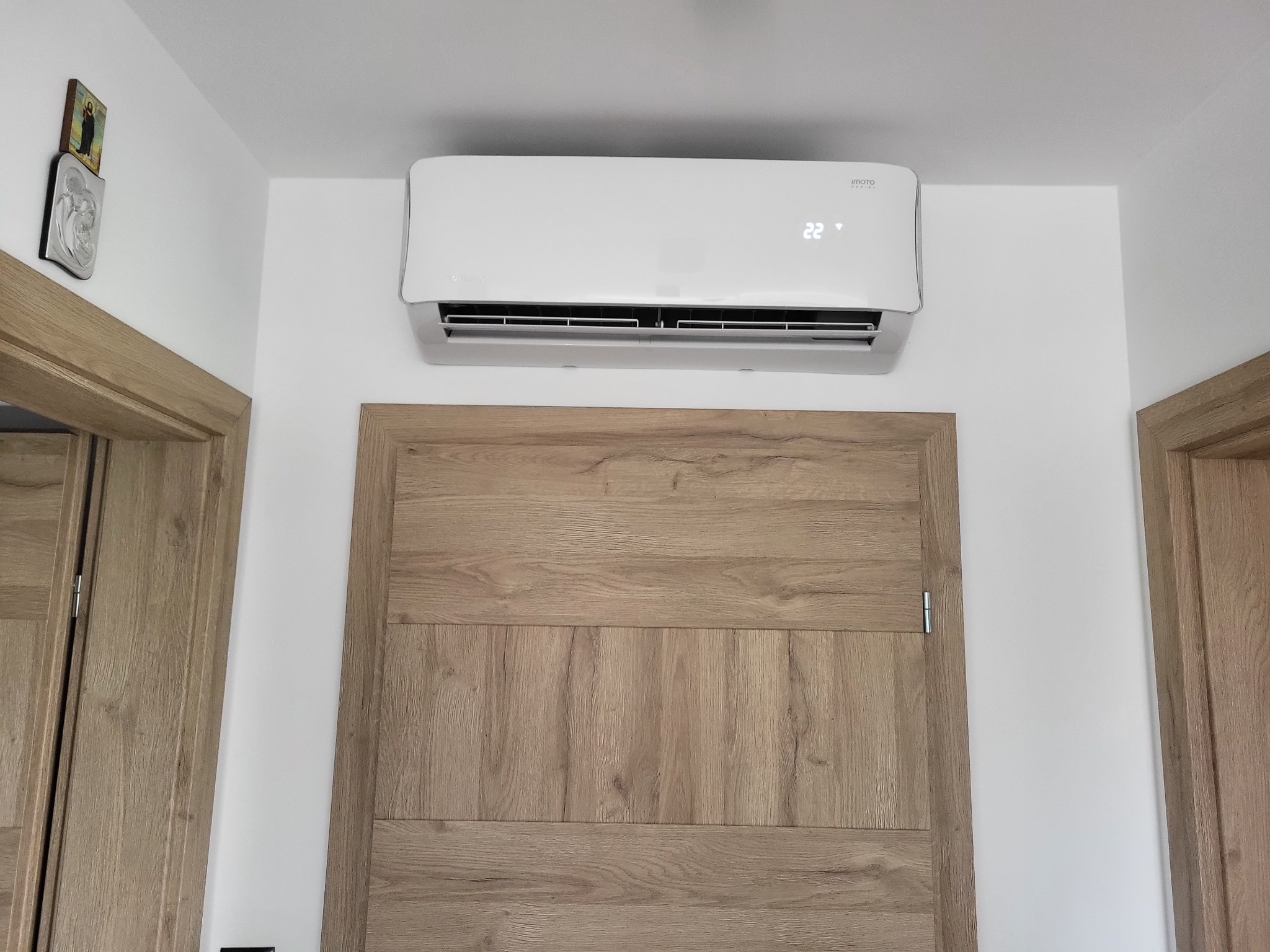 Biały klimatyzator ścienny z wyświetlaczem temperatury 22 stopnie zamontowany nad drewnianymi drzwiami w jasnym pomieszczeniu, widoczna ikona religijna na ścianie.