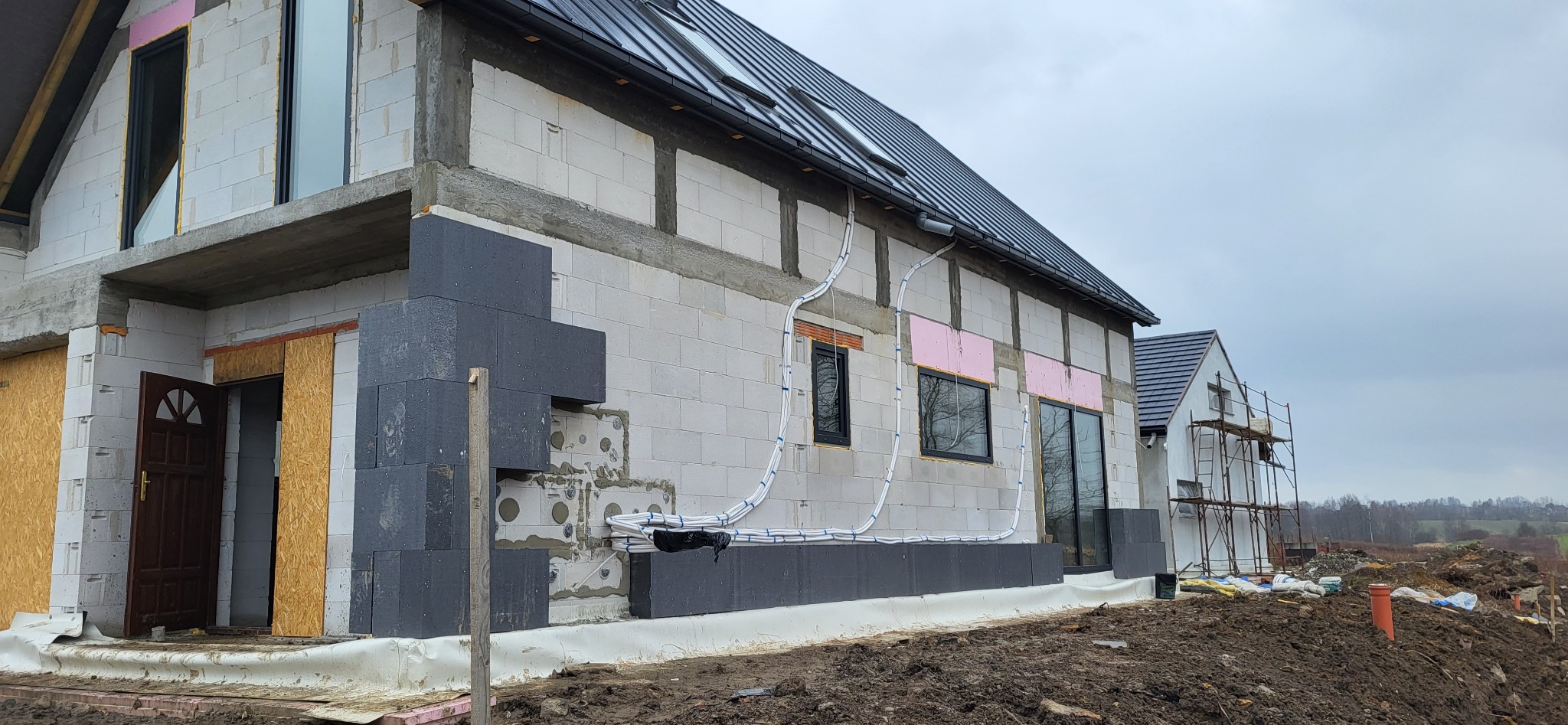 Surowy stan budynku z białego bloczka, ocieplonego częściowo szarym styropianem, widoczne rury wentylacyjne na elewacji, okna z czarnymi ramami, dach z ciemnej blachy, w tle drugi budynek w trakcie...
