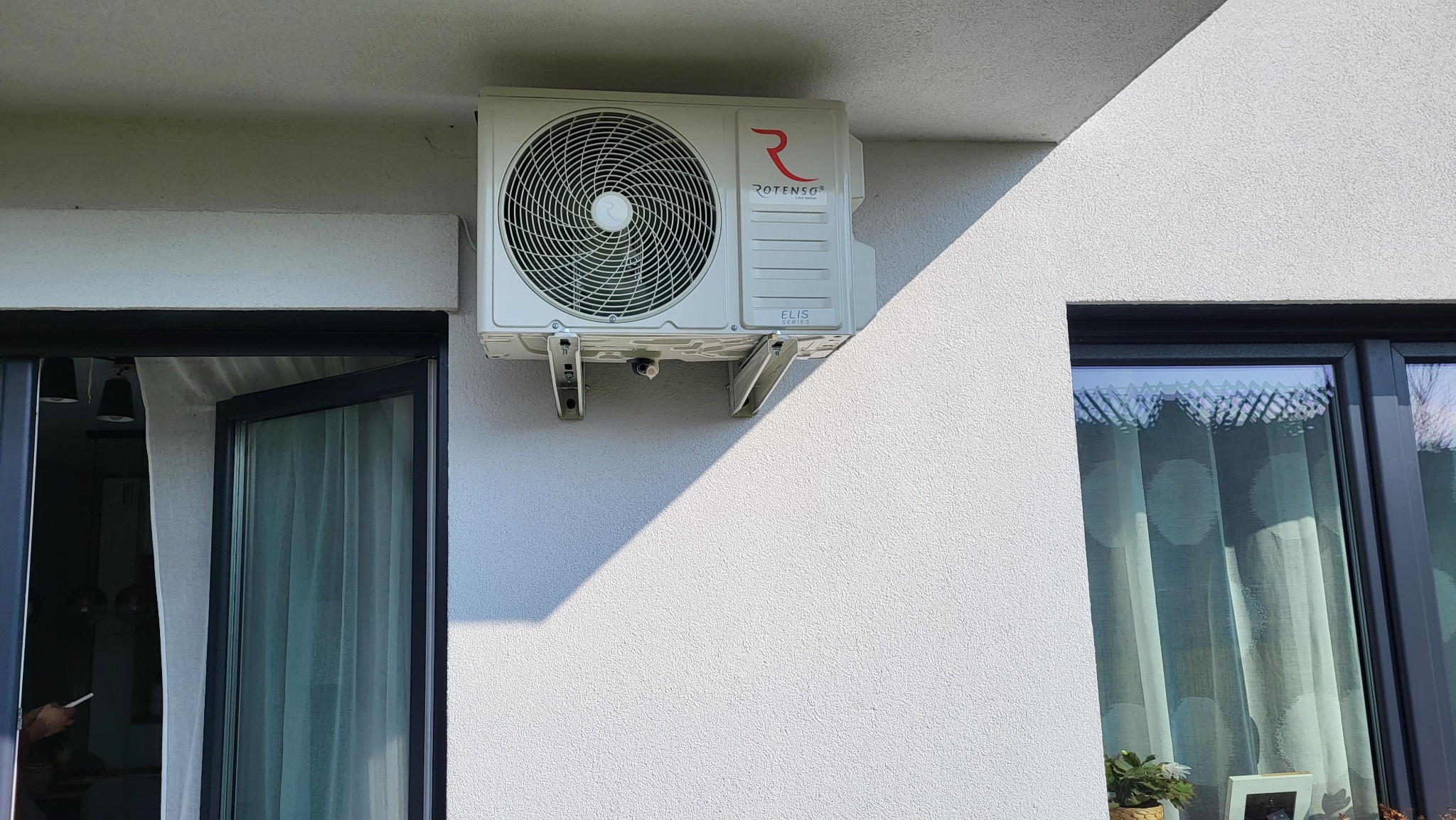 Zewnętrzny klimatyzator Rotenso ELIS Series zamontowany na białej, teksturowanej elewacji budynku, widoczny z boku okna z otwartymi drzwiami balkonowymi.