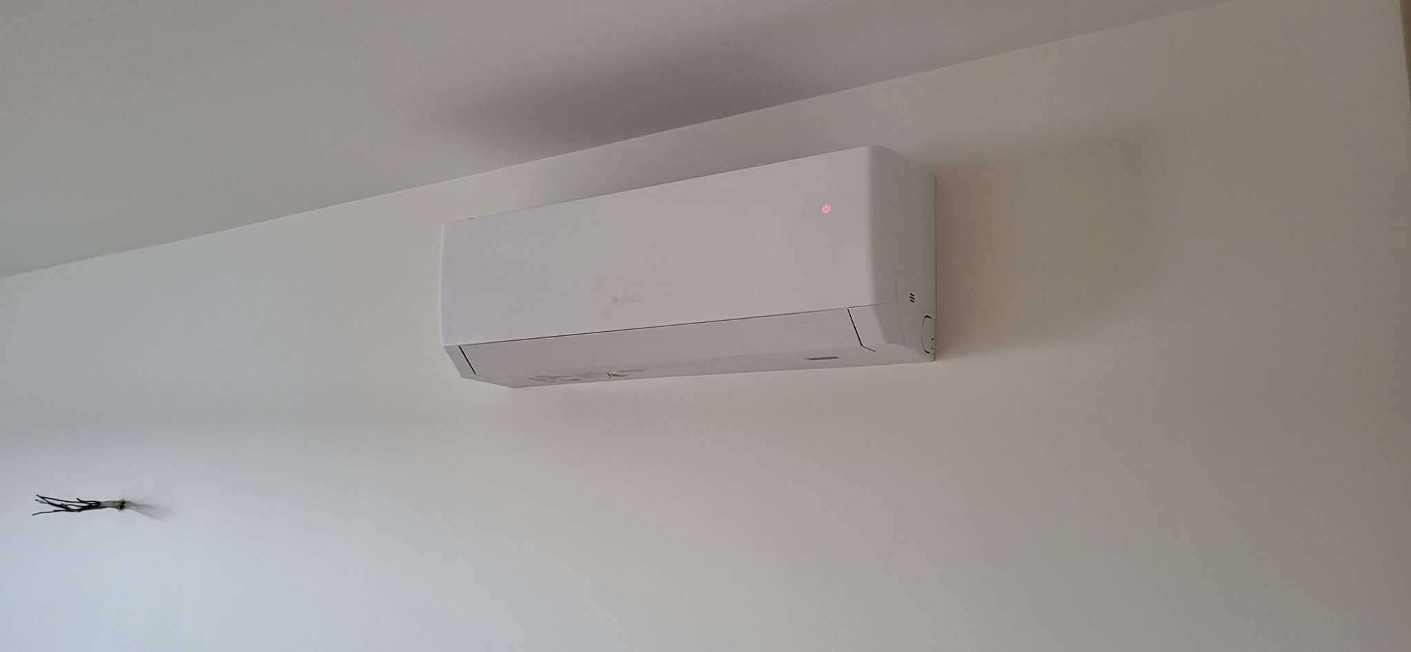 Biały klimatyzator marki Gree zamontowany na jasnej ścianie, widoczny czerwony wskaźnik zasilania i ślady po instalacji elektrycznej.