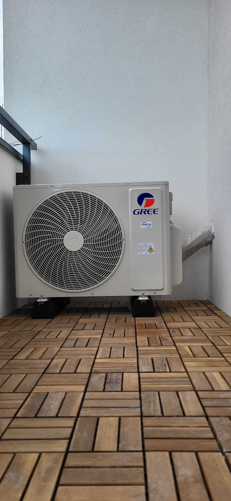 Zewnętrzna jednostka klimatyzacji marki Gree, zamontowana na balkonie wyłożonym drewnianą podłogą, z widocznymi czarnymi podkładkami antywibracyjnymi.