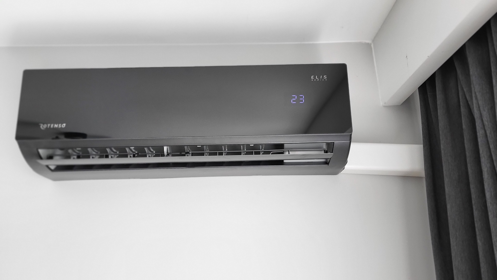 Czarny klimatyzator Rotenso Elis Series zamontowany na białej ścianie, wyświetlający temperaturę 23 stopnie, z widoczną częścią szarej zasłony po prawej stronie.