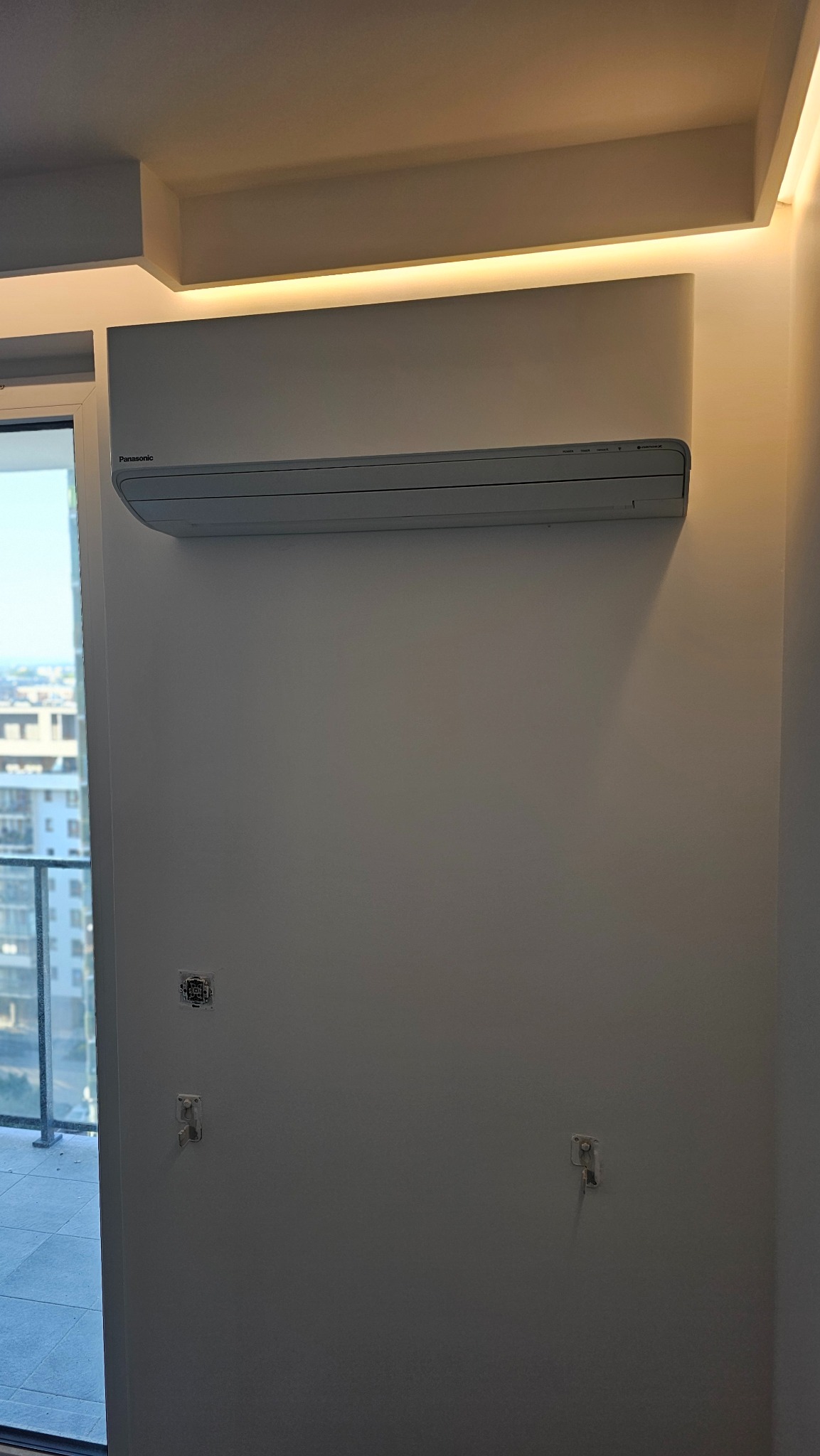 Zamontowana jednostka klimatyzacyjna Panasonic na białej ścianie w nowoczesnym wnętrzu z widocznymi elementami instalacji elektrycznej i wyjściem na balkon z widokiem na miasto.