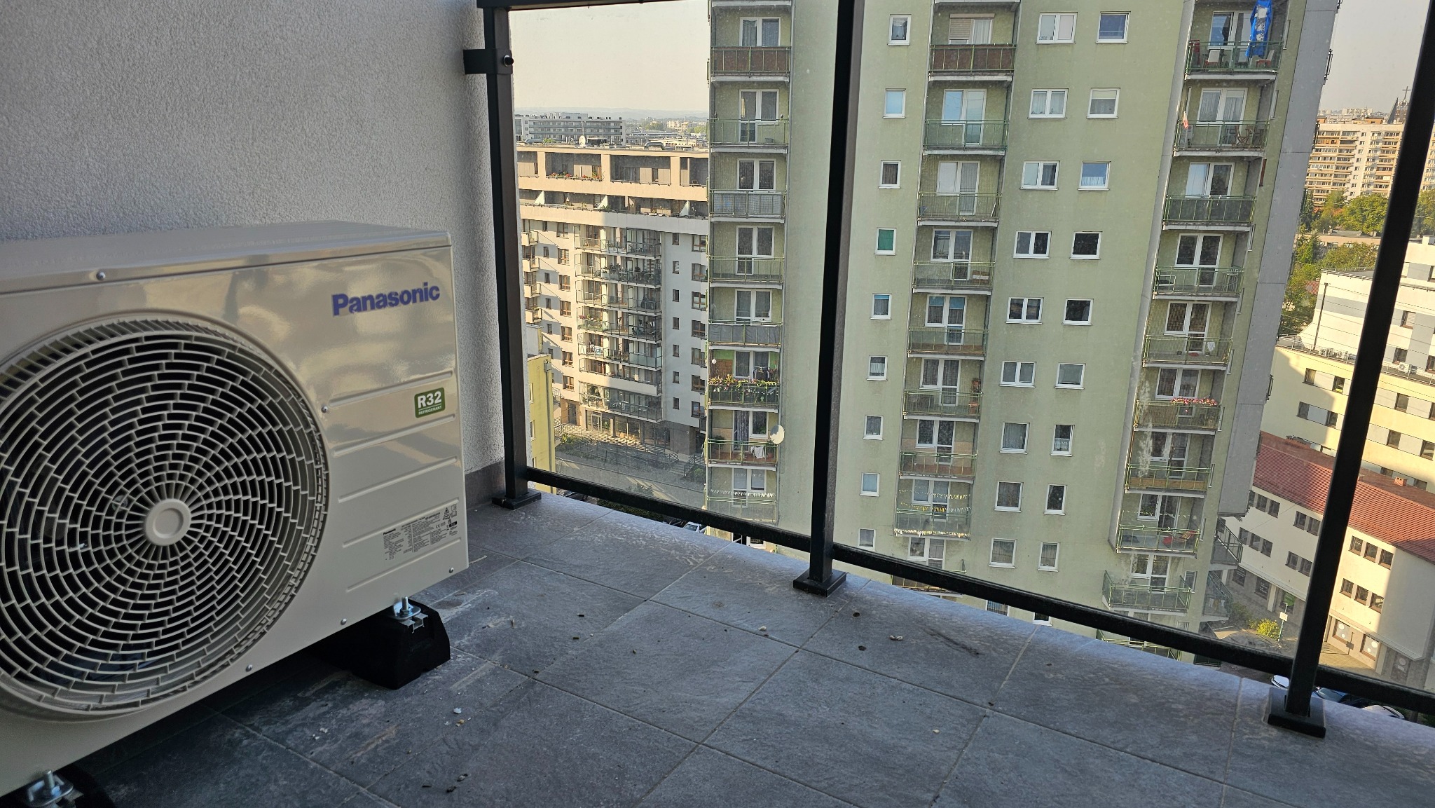 Jednostka zewnętrzna klimatyzatora Panasonic zamontowana na balkonie z widokiem na budynki mieszkalne w tle.