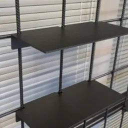 Metalowa konstrukcja regału z czarnymi półkami i prętami o skręconej fakturze na tle białych żaluzji.