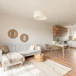 Jasne, przestronne wnętrze: salon z szarą sofą narożną, drewnianym stolikiem kawowym, dywanem w paski i okrągłymi lustrami na ścianie, przechodzące w jadalnię z drewnianym stołem i plecionymi...