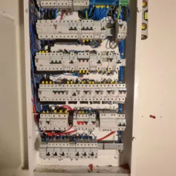 Otwarta skrzynka elektryczna z licznymi bezpiecznikami, przewodami w różnych kolorach i poziomica przymocowana z boku, dodatkowa obudowa na dole.