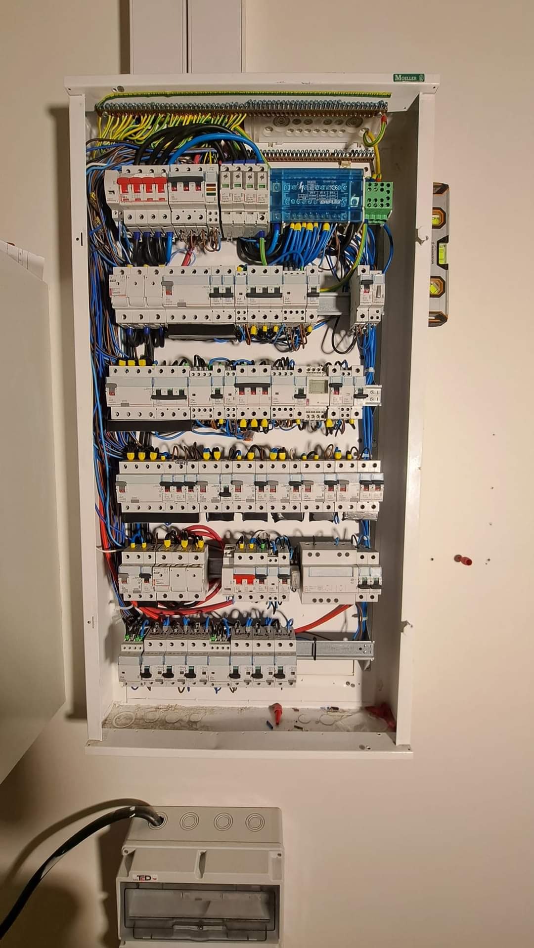 Otwarta skrzynka elektryczna z licznymi bezpiecznikami, przewodami w różnych kolorach i poziomica przymocowana z boku, dodatkowa obudowa na dole.