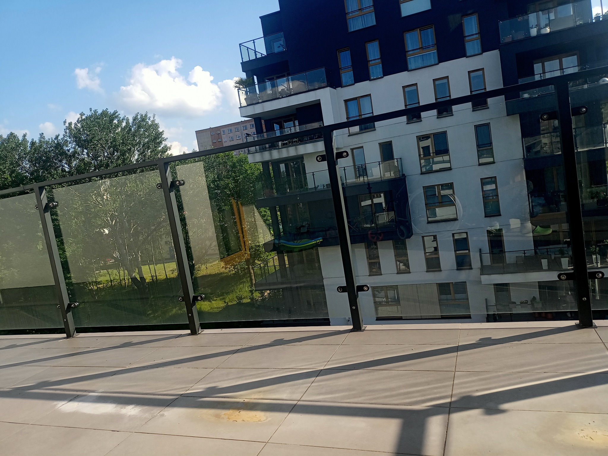 Widok z balkonu na nowoczesny budynek mieszkalny z przeszklonymi balustradami, cień barierki na jasnych płytkach podłogowych.