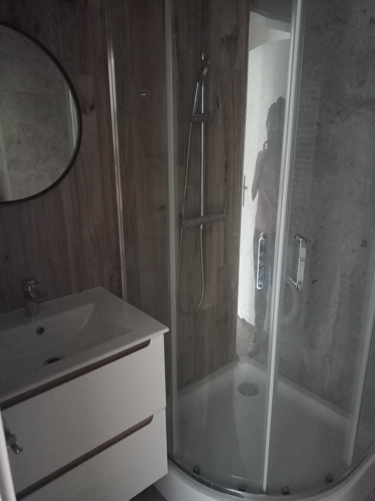 Nowoczesna łazienka z kabiną prysznicową z przezroczystego szkła, białą szafką pod umywalką i okrągłym lustrem na ścianie z drewnianą okładziną.