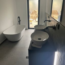Usługi leśne - Nowoczesna łazienka w trakcie wykańczania, z wolnostojącą białą wanną, podwójną umywalką nablatową na czarnym blacie i czarną armaturą; widok przez okno na plac budowy z hałdą ziemi.