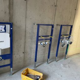 Usługi leśne - Stelaże podtynkowe Geberit zamontowane na surowej ścianie z betonu, widoczny laser krzyżowy wyznaczający poziom, żółty pojemnik z narzędziami na podłodze.