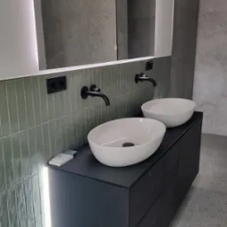 Nowoczesna łazienka z dwoma białymi umywalkami nablatowymi na czarnej szafce, podświetlane lustro z ledowym oświetleniem, zielone pionowe płytki na ścianie i czarne baterie.