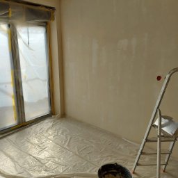 Usługi leśne - Pokój w trakcie remontu: ściany przygotowane do malowania, podłoga i okno zabezpieczone folią, drabina i wiadro z zaprawą w pomieszczeniu.