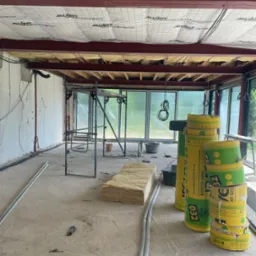 Wnętrze budynku w trakcie ocieplania: widoczne stalowe belki stropowe z izolacją, rusztowanie, rolki wełny mineralnej w folii, przewody elektryczne i surowe ściany.